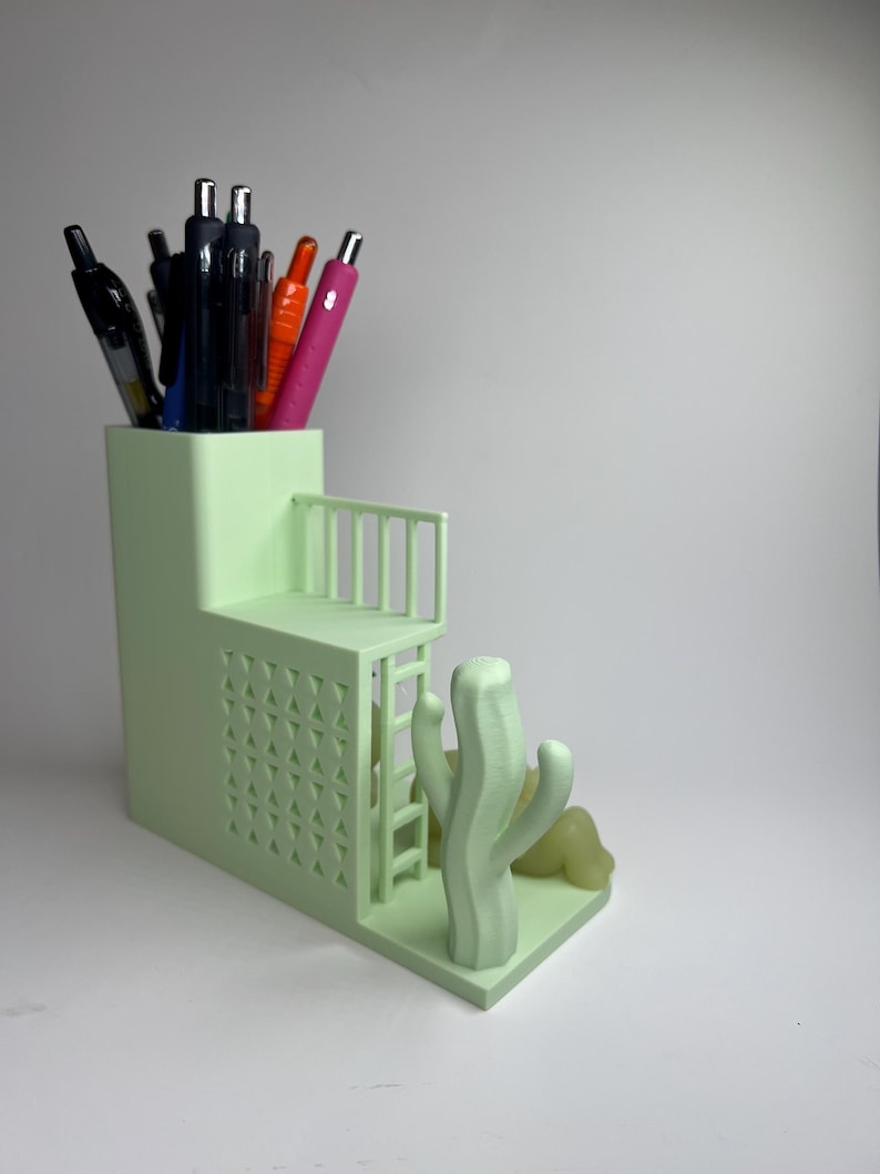 May include: A light green desk organizer with a tiered design, featuring a small balcony and a cactus sculpture. The organizer holds various pens and pencils. The design includes a ladder and geometric cutouts.