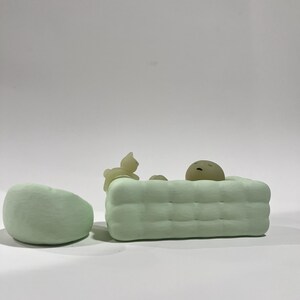 Puede incluir: Un conjunto de esculturas minimalistas de color verde claro que presenta una silla redondeada y una forma rectangular con un patr&oacute;n acolchado, posiblemente una cama o un sof&aacute;. Peque&ntilde;as figuras abstractas de un gato y una cara durmiente.