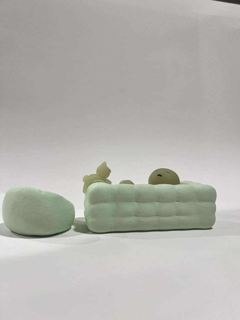 Puede incluir: Un conjunto de esculturas minimalistas de color verde claro que presenta una silla redondeada y una forma rectangular con un patr&oacute;n acolchado, posiblemente una cama o un sof&aacute;. Peque&ntilde;as figuras abstractas de un gato y una cara durmiente.