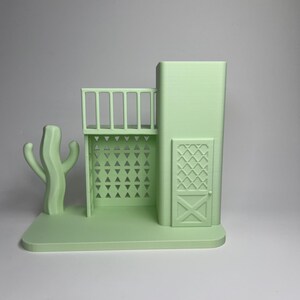 May include: A light green 3D-printed model of a Southwestern-style building with a balcony, door, and a cactus. The building has a rectangular base and features geometric cutouts and a decorative door. The cactus is on the left side.