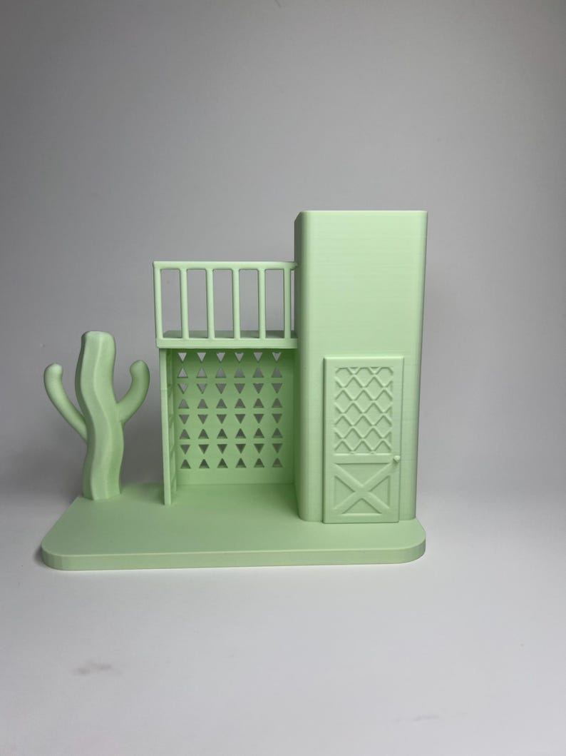 May include: A light green 3D-printed model of a Southwestern-style building with a balcony, door, and a cactus. The building has a rectangular base and features geometric cutouts and a decorative door. The cactus is on the left side.