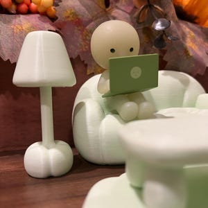 May include: A miniature scene featuring a light green figurine seated on a sofa, using a laptop. A small lamp and table are also present. The background includes fall foliage with orange, yellow, and brown tones. The laptop screen displays a small face.