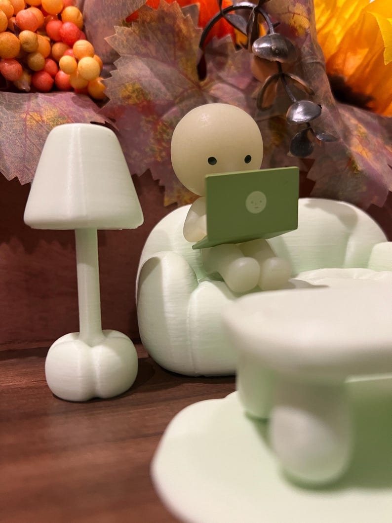 May include: A miniature scene featuring a light green figurine seated on a sofa, using a laptop. A small lamp and table are also present. The background includes fall foliage with orange, yellow, and brown tones. The laptop screen displays a small face.