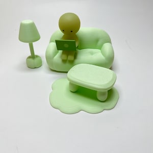 May include: A light green miniature room scene featuring a small couch, a table, a lamp, and a figure with a laptop. The figure is sitting on the couch, working on the laptop. The lamp is next to the couch. The table is in front of the couch.