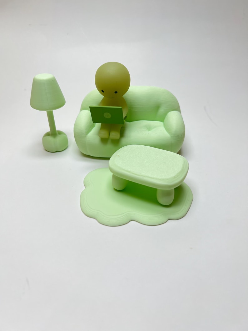 May include: A light green miniature room scene featuring a small couch, a table, a lamp, and a figure with a laptop. The figure is sitting on the couch, working on the laptop. The lamp is next to the couch. The table is in front of the couch.