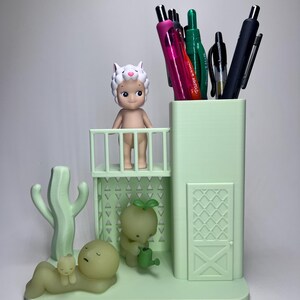 May include: A light green desk organizer featuring a pen holder, a small balcony with a figurine, and a cactus. The organizer also includes a small figurine with a watering can and a figurine lying down with a smaller figurine on top. Pens are in the pen holder.