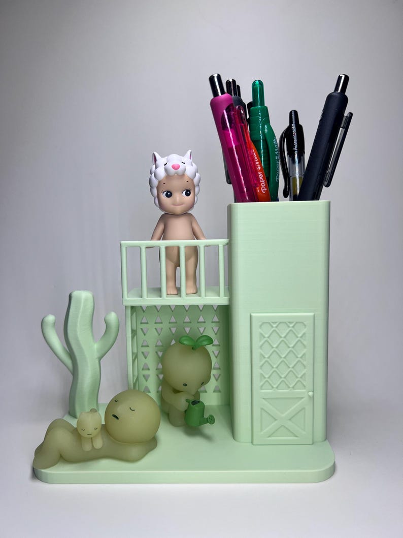 May include: A light green desk organizer featuring a pen holder, a small balcony with a figurine, and a cactus. The organizer also includes a small figurine with a watering can and a figurine lying down with a smaller figurine on top. Pens are in the pen holder.