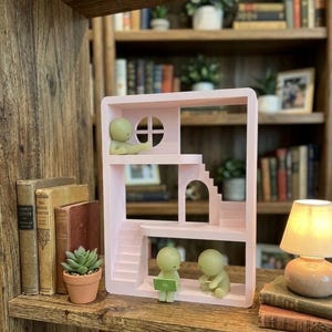 May include: A light pink dollhouse-style shelf with miniature figures. The shelf has stairs, a window, and a small archway. Two figures are seated, one with a laptop. A small potted succulent and vintage books are on the shelf.