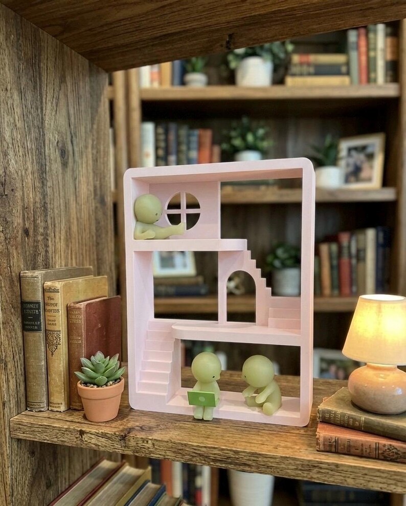 May include: A light pink dollhouse-style shelf with miniature figures. The shelf has stairs, a window, and a small archway. Two figures are seated, one with a laptop. A small potted succulent and vintage books are on the shelf.