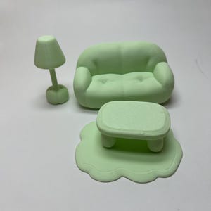 May include: A miniature mint green living room set, including a sofa, coffee table on a rug, and a lamp. The furniture has rounded edges and a smooth texture, creating a playful and whimsical aesthetic. The set is designed for a dollhouse or similar decorative use.