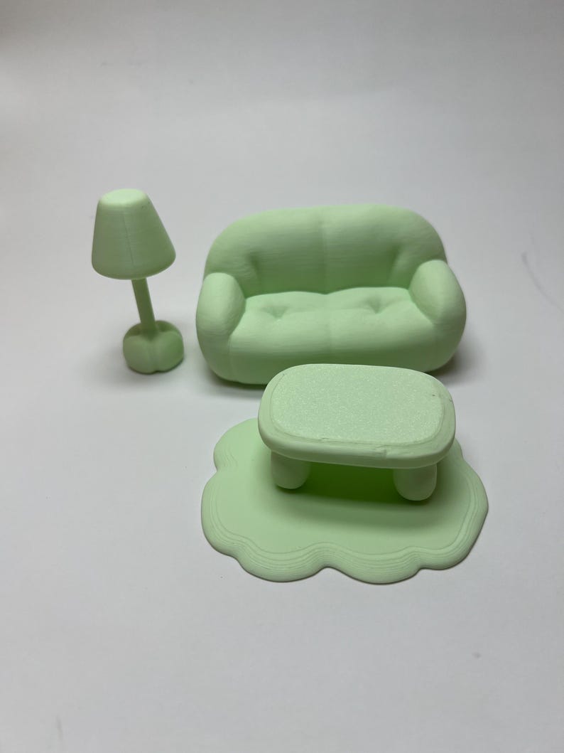 May include: A miniature mint green living room set, including a sofa, coffee table on a rug, and a lamp. The furniture has rounded edges and a smooth texture, creating a playful and whimsical aesthetic. The set is designed for a dollhouse or similar decorative use.