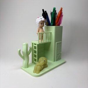 May include: A light green pen holder with a cactus and two figurines. The pen holder has a small balcony with a figurine wearing a sheep hat. Several pens are inside the holder. Another figurine is lying down.