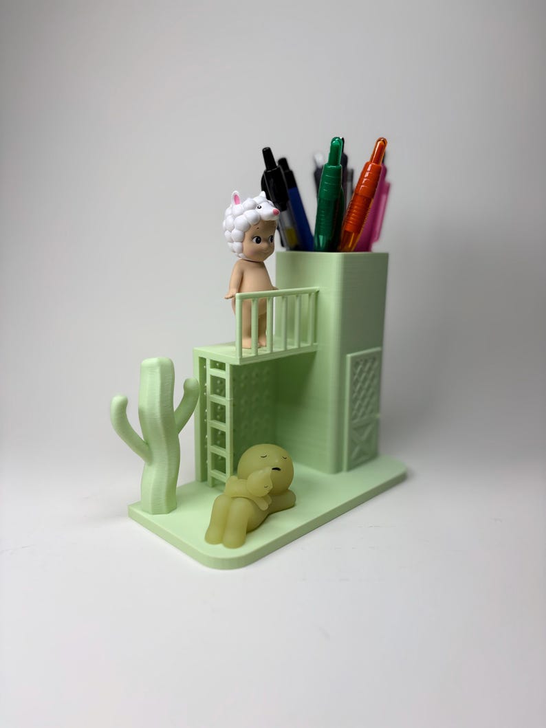 May include: A light green pen holder with a cactus and two figurines. The pen holder has a small balcony with a figurine wearing a sheep hat. Several pens are inside the holder. Another figurine is lying down.