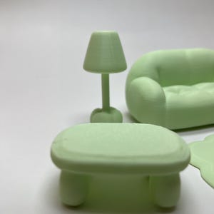 May include: A miniature light green living room set, including a small table, a lamp, and a rounded sofa. The table has rounded legs, and the lamp has a conical shade. The sofa has a curved design.