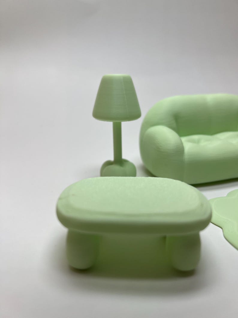 May include: A miniature light green living room set, including a small table, a lamp, and a rounded sofa. The table has rounded legs, and the lamp has a conical shade. The sofa has a curved design.