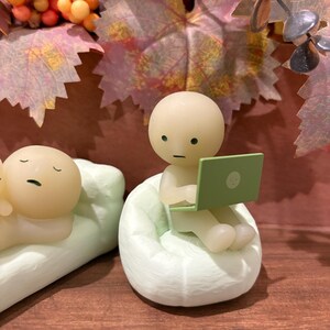 Puede incluir: Una figura de una persona sentada en un puf verde claro, usando una computadora port&aacute;til verde. Otra figura est&aacute; acostada en una almohada verde claro. El fondo incluye follaje de oto&ntilde;o y una superficie de madera.