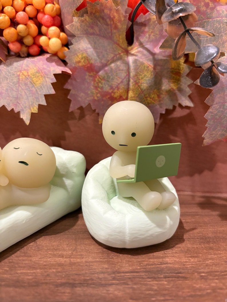 Puede incluir: Una figura de una persona sentada en un puf verde claro, usando una computadora port&aacute;til verde. Otra figura est&aacute; acostada en una almohada verde claro. El fondo incluye follaje de oto&ntilde;o y una superficie de madera.