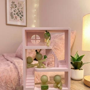 May include: A pink dollhouse-style shelf unit with multiple levels and miniature figures. The figures are light green and appear to be interacting with each other. The shelf has a window and stairs. A framed botanical print hangs on the wall.