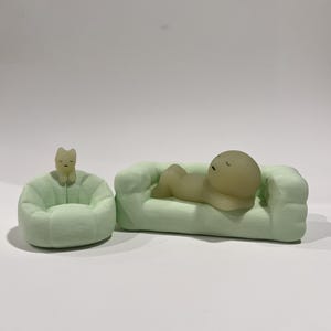 Puede incluir: Un sof&aacute; en miniatura verde claro con una figura acostada sobre &eacute;l, junto a un puf a juego con una figura de gato. Las figuras son de color amarillo p&aacute;lido. Los objetos est&aacute;n sobre una superficie blanca.