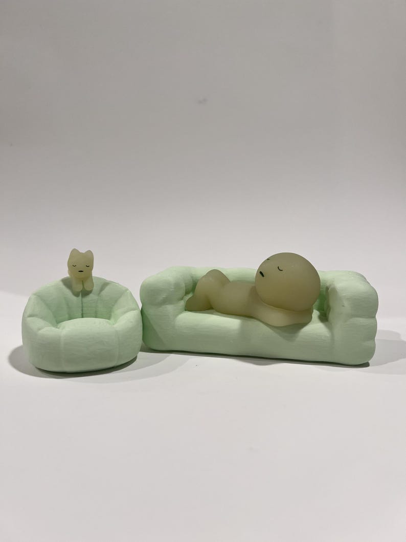 Puede incluir: Un sof&aacute; en miniatura verde claro con una figura acostada sobre &eacute;l, junto a un puf a juego con una figura de gato. Las figuras son de color amarillo p&aacute;lido. Los objetos est&aacute;n sobre una superficie blanca.