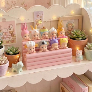 Könnte beinhalten: Eine Präsentation von Sammlerfiguren mit Tierhut-Motiven auf einem rosa Regal. Das Regal ist mit kleinen Sukkulenten in Töpfen, Lichterketten und Miniatur-Tierfiguren dekoriert. Im Hintergrund befindet sich eine florale Tapete.