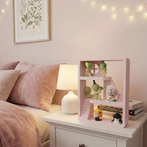 May include: A pink, tiered display shelf with miniature figurines. The shelf is on a white nightstand next to a bed with pink bedding. A botanical print hangs on the wall above the bed. String lights are draped across the wall.