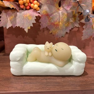 Puede incluir: Un sof&aacute; en miniatura verde claro con una figura durmiente y una peque&ntilde;a figura de gato. La figura es de color beige claro con los ojos cerrados, y el gato es de color beige claro. El sof&aacute; est&aacute; sobre una superficie marr&oacute;n oscuro.