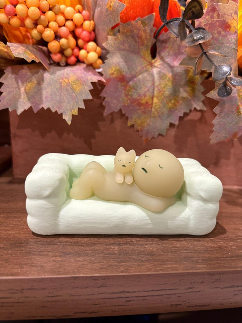 Puede incluir: Un sof&aacute; en miniatura verde claro con una figura durmiente y una peque&ntilde;a figura de gato. La figura es de color beige claro con los ojos cerrados, y el gato es de color beige claro. El sof&aacute; est&aacute; sobre una superficie marr&oacute;n oscuro.