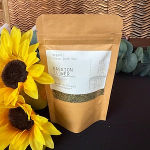 May include: A brown paper bag of organic loose leaf tea labeled "PASSION FLOWER" with the brand name "Earth & Thread". The bag is next to bright yellow sunflowers with dark brown centers. The background is a dark brown.