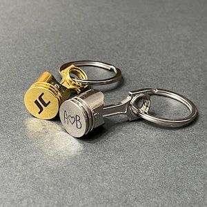 May include: Three metal piston keyrings. One is gold-coloured with the initials "JC", another is silver-coloured with the initials "A♡B". Each keyring has a ring for attaching keys. The keyrings are on a grey surface.