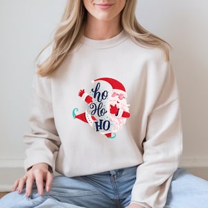 May include: A cream-colored sweatshirt featuring a cartoon Santa Claus design. The Santa illustration has a red hat, white beard, and sunglasses, with the words "ho ho ho" in a playful font. The design is centered on the front of the sweatshirt.