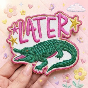 May include: Embroidered patch featuring a green alligator with an open mouth, the word "LATER" in pink, and star accents. The patch has a white background and a pink border. The background is light pink with embroidered flowers, hearts, butterflies, and stars.