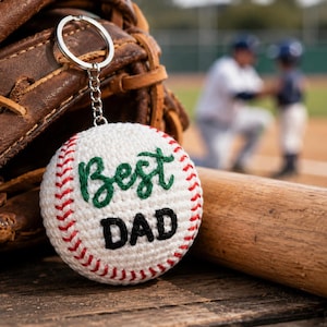 Puede incluir: Un llavero de béisbol blanco con costuras rojas y las palabras "Best DAD" en verde y negro. El llavero está delante de un guante de béisbol y un bate de béisbol de madera.