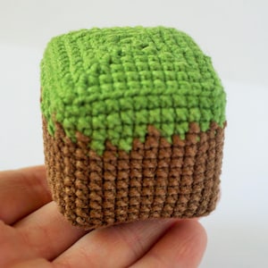 May include: A crocheted cube toy with a green top representing grass and a brown bottom. The handmade block is held in a hand and is a Minecraft-inspired design. The cube measures approximately 6.35 cm.