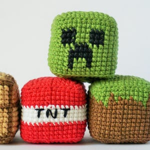 May include: A collection of crocheted Minecraft-inspired cubes. The set includes a green Creeper face, a red TNT block with "TNT" text, a brown and tan treasure chest, and a grass block. These handmade items are ideal for fans of the popular video game.