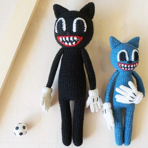 May include: Two crocheted cartoon cat dolls, one black and one blue, with white gloves and large, toothy grins. A small soccer ball sits nearby. The dolls have large eyes and are standing upright.