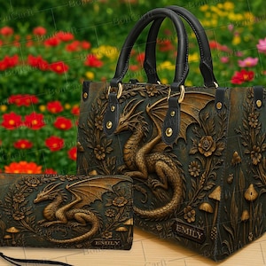 May include: A dark green handbag and matching wallet set with a dragon design. The dragon is bronze-colored and has detailed wings. The bag has black handles and gold-colored hardware. The wallet has a wrist strap and the name "EMILY" is visible on both items.