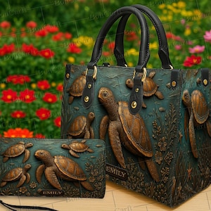 Puede incluir: Un juego de bolso y cartera de color verde azulado con tortugas marinas en relieve. El bolso tiene asas negras y herrajes dorados. La cartera tiene una correa de muñeca y el nombre "EMILY" es visible en ambos artículos. El fondo es una escena floral borrosa.