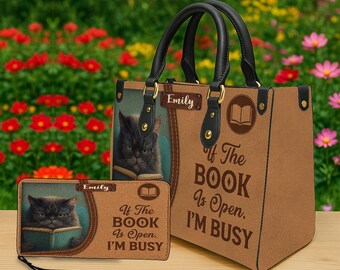Puede incluir: Un bolso tote marrón y una cartera a juego con un gato con gafas leyendo un libro. El bolso y la cartera tienen el texto "If The BOOK Is Open. I'M BUSY". El bolso tiene asas negras y herrajes dorados.
