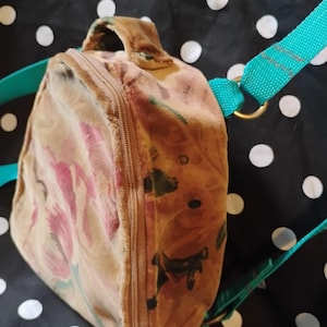 May include: A small, vintage-style backpack with a floral pattern in shades of pink, brown, and green. The bag has a zippered closure, a top handle, and teal-colored straps. The background is black with white polka dots.
