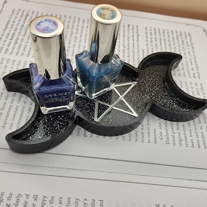 Pode incluir: Um suporte de verniz de unhas preto, com glitter, num design de lua crescente e pentagrama, segurando dois frascos de verniz de unhas. Os frascos de verniz têm tampas prateadas e são preenchidos com verniz azul e roxo.