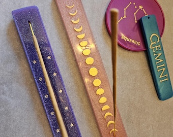 Puede incluir: Un conjunto de porta inciensos y un posavasos con temática zodiacal. Los soportes rectangulares son morados y rosas, adornados con lunas y estrellas doradas. También hay una etiqueta Géminis turquesa y un posavasos Acuario morado con detalles de constelaciones doradas.