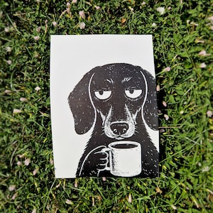 May include: A black and white print on paper featuring a stylized illustration of a dog with long ears and a tired expression, holding a white coffee mug with both paws. The dog is depicted in a graphic, high-contrast ink style on a white background.