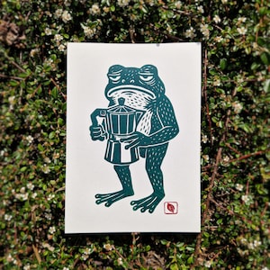 Könnte beinhalten: Ein türkisgrüner Druck einer Froschfigur, die einen Kaffeezubereiter hält. Der Frosch hat einen mürrischen Gesichtsausdruck und steht aufrecht. Ein rotes Blatt befindet sich in der Ecke. Der Druck befindet sich auf weißem Hintergrund.