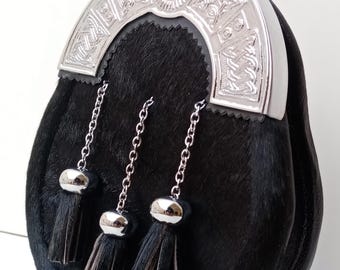 Op de afbeelding: Een zwarte leren sporran met een zilveren cantle met Keltische knoopwerk. Het heeft drie hangende kwasten met zilveren doppen en kettingen. Een traditioneel accessoire.