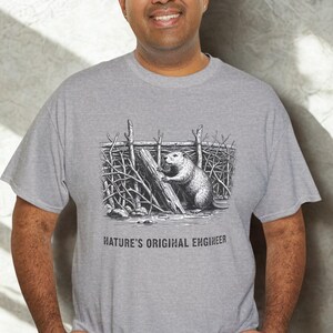 May include: A grey t-shirt with a black and white illustration of a beaver by a dam. The text "NATURE'S ORIGINAL ENGINEER" is printed below the graphic. The shirt is made of a soft material. The person is wearing the shirt.