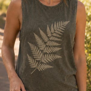 Puede incluir: Una camiseta sin mangas de color verde oliva lavado que presenta un gran gráfico de hoja de helecho de color claro impreso en la parte delantera. La prenda tiene cuello redondo, sisas anchas y un corte relajado con textura desgastada.