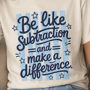 May include: A cream-colored short-sleeve t-shirt featuring a graphic design with light blue vertical stripes and dark blue text that reads, Be like Subtraction and make a difference, accented with small star shapes surrounding the lettering.