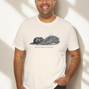 May include: Cream-coloured t-shirt with a black and white illustration of a beaver and a pile of logs. The text "Nature's Original Engineer" is printed below the image. The shirt is being worn by a person.