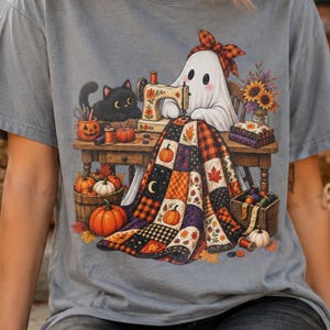May include: A grey short-sleeve t-shirt featuring a graphic of a white ghost wearing a plaid headband, sitting at a wooden sewing machine. The ghost is sewing a colourful patchwork quilt decorated with pumpkins, leaves, and a black cat nearby.